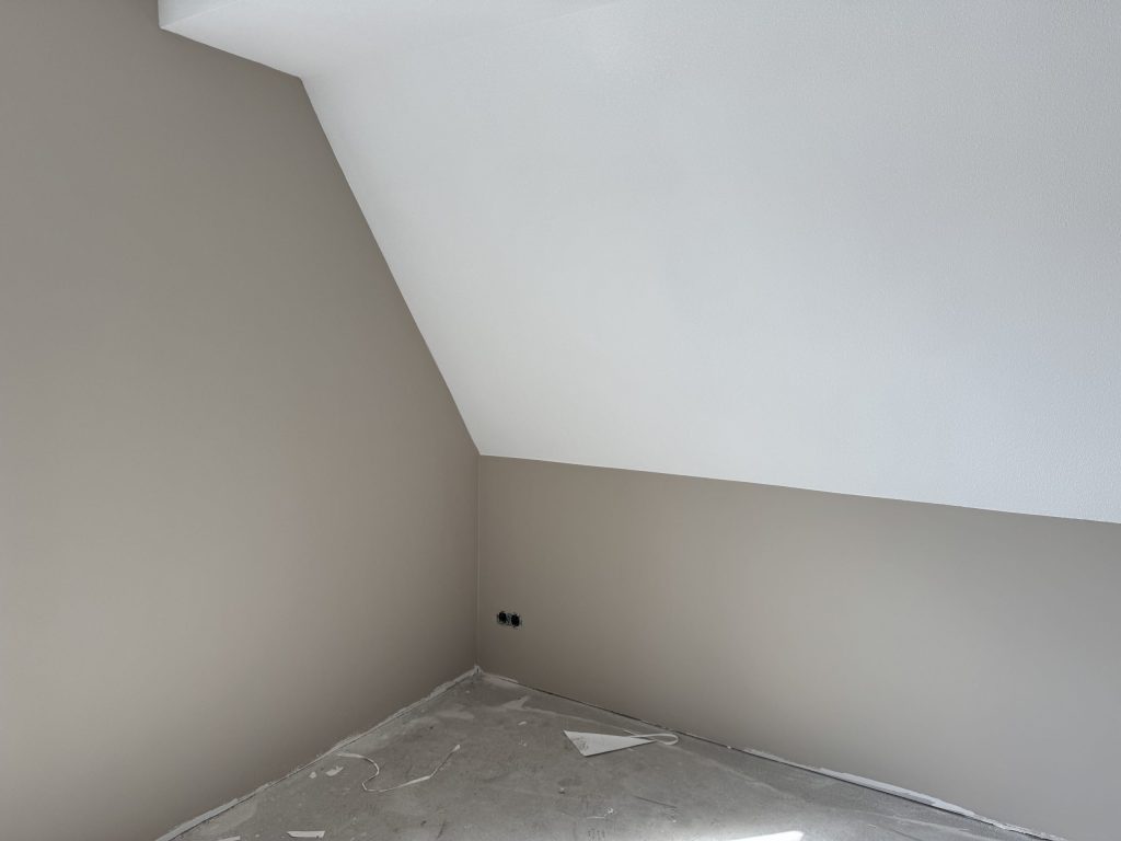Zolderkamer met een witte schuine wand boven taupe gesausde muren, net na het behangen